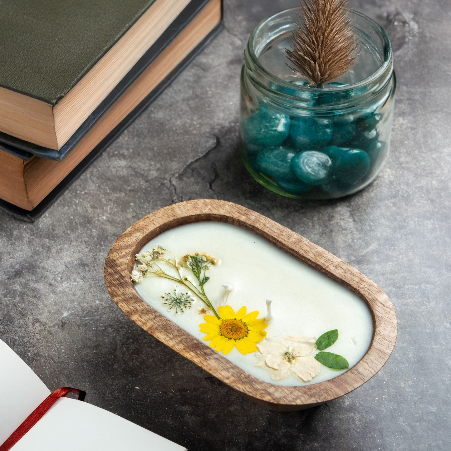 Candle with pressed flowers in a wooden holder on a gray surface with books and a jar.