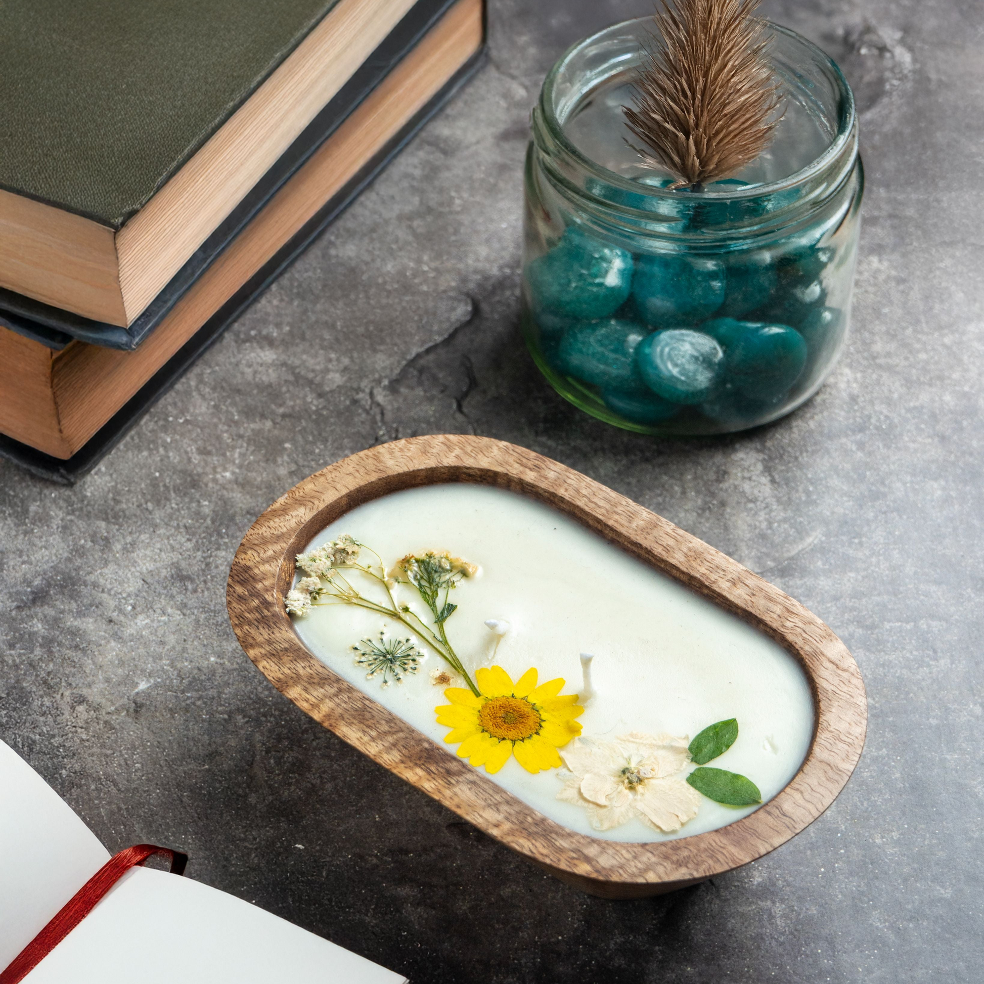 Candle with pressed flowers in a wooden holder on a gray surface with books and a jar.