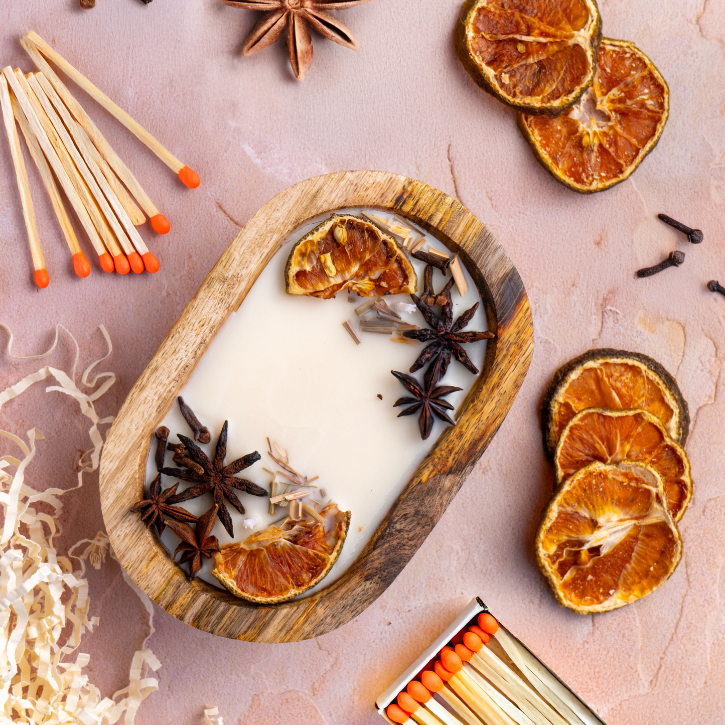 Wooden tray with lit candle, dried oranges, star anise, and matches on a pink background