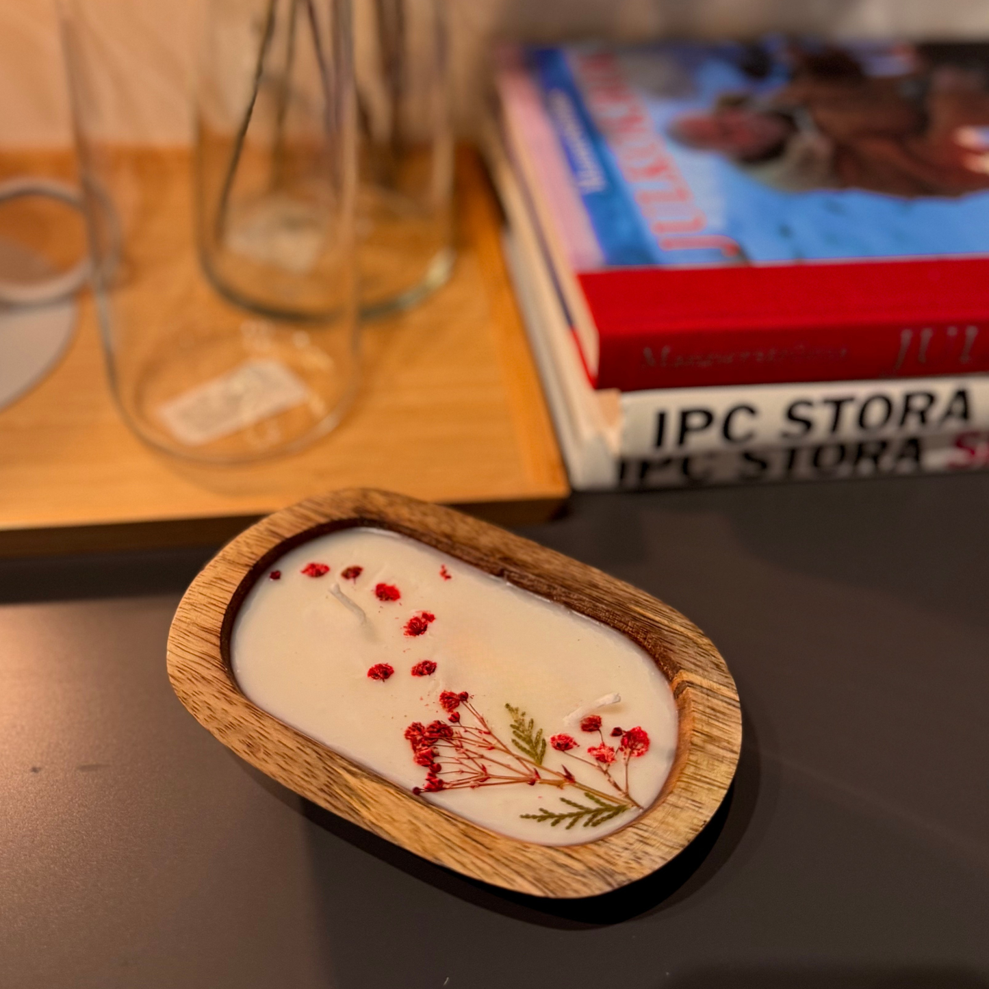 Candle with dried flowers in a wooden holder on a surface with books and glasses in the background.