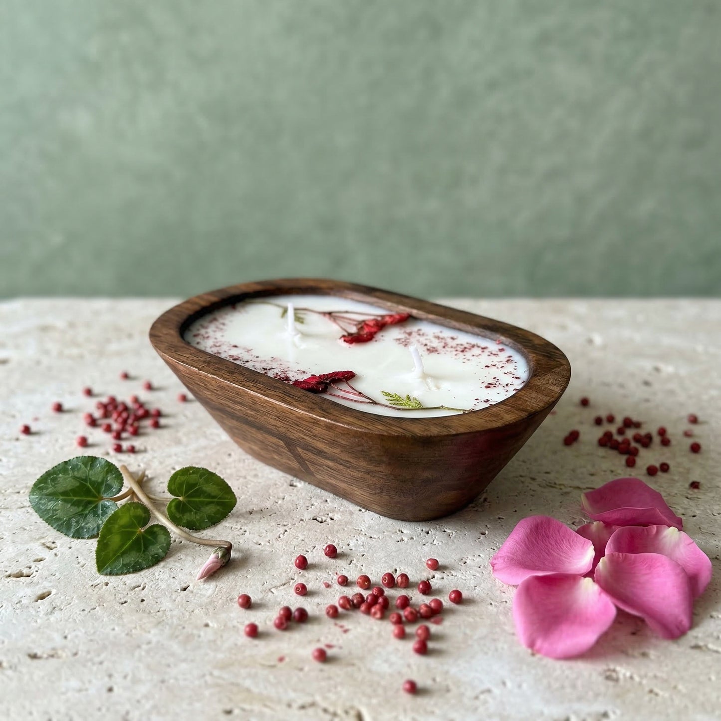 Wooden bowl candle with a white substance, red berries, and pink petals on a light surface with a green background