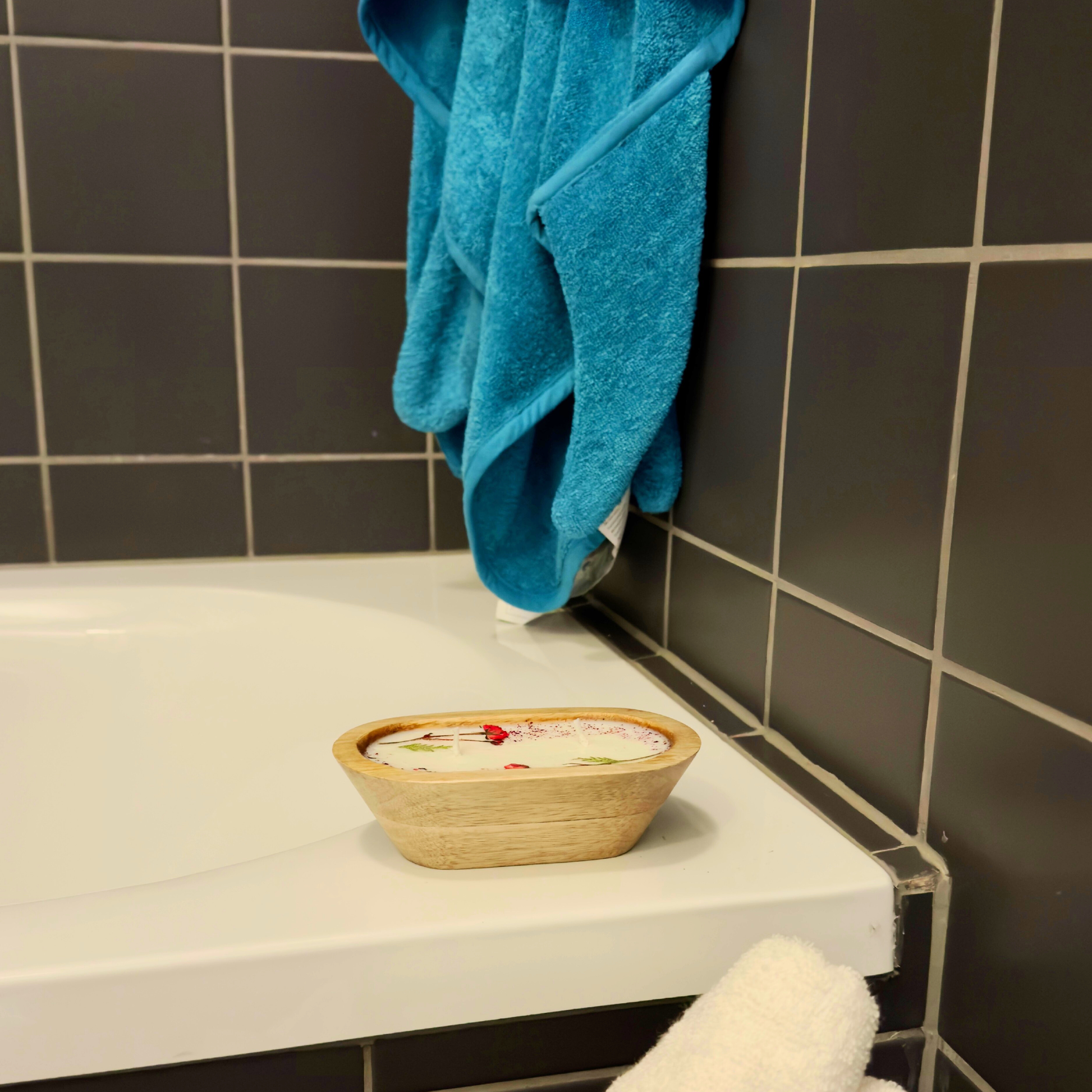 Wooden bowl candle with decorative items on a bathroom counter next to a blue towel.