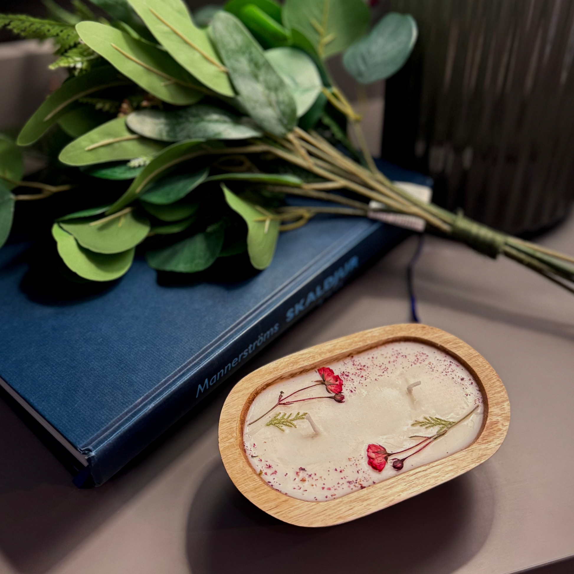 Wooden bowl candle with floral design on a surface with green leaves and a blue book in the background