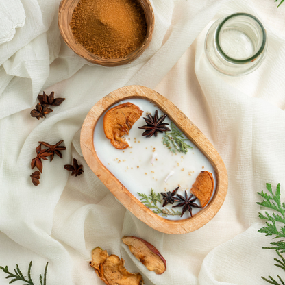 Spiced candle in a wooden bowl with cinnamon, star anise, and other spices on a white fabric background.