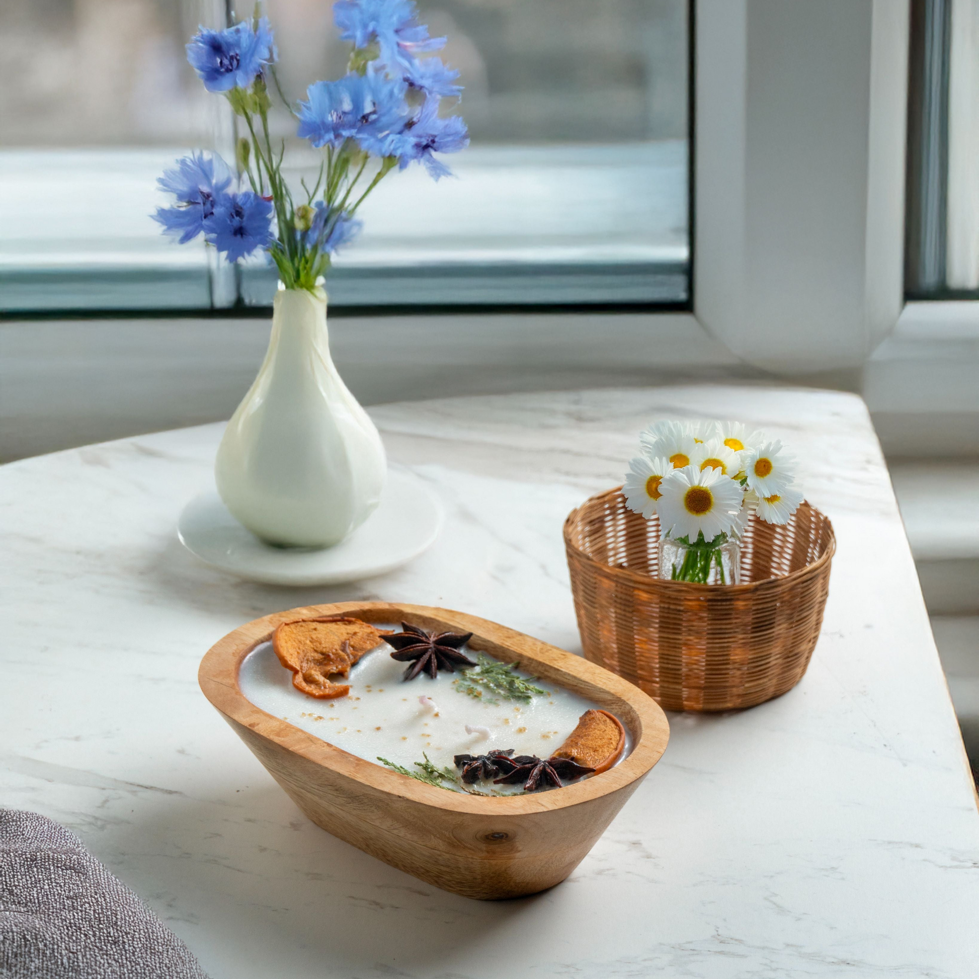 Wooden bowl with decorative items on a marble surface, next to a vase with blue flowers and a basket with white flowers.