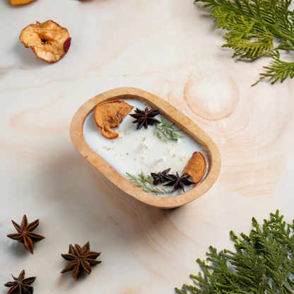 Wooden bowl with a candle, star anise, and cinnamon on a marble surface with greenery.