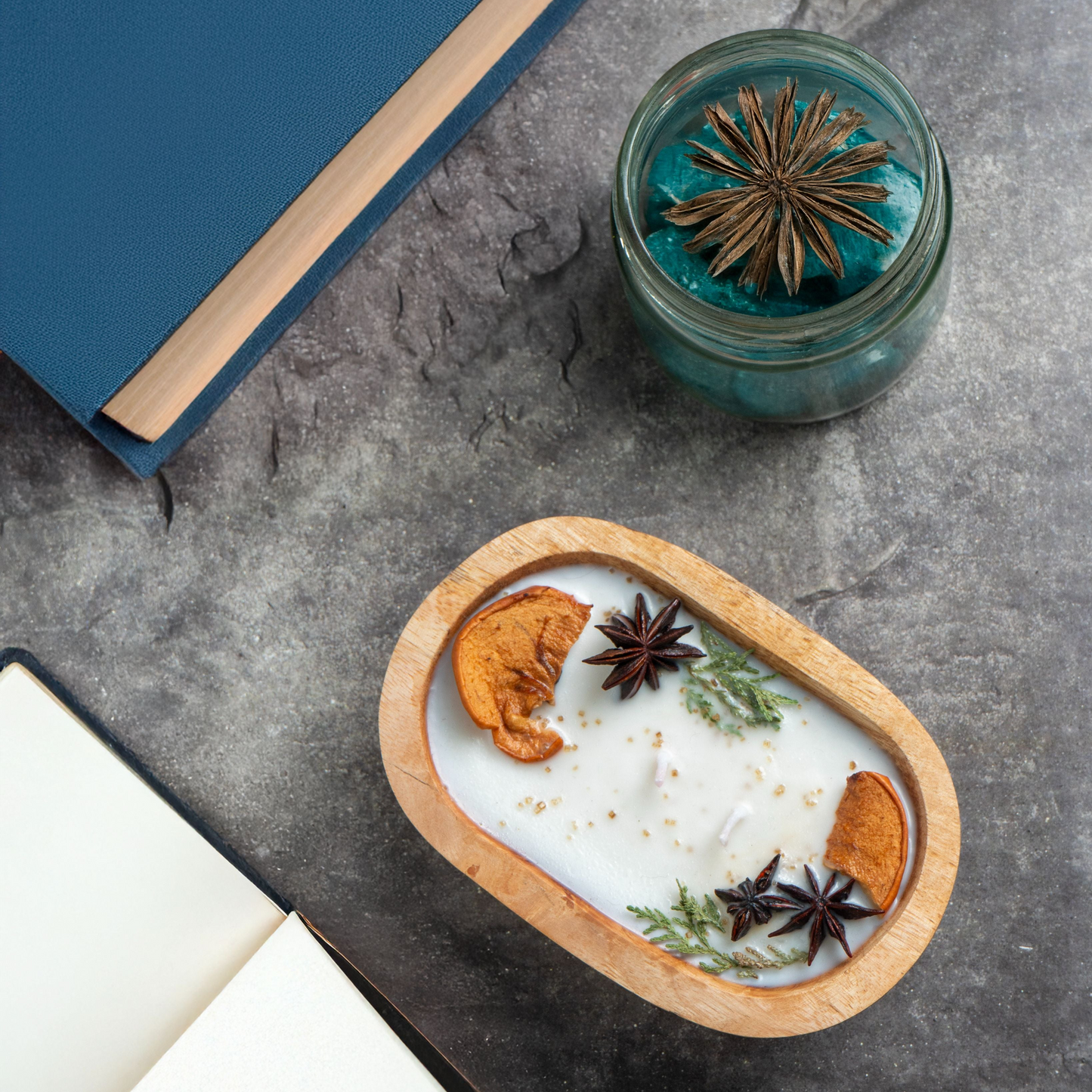 Candle with natural elements in a wooden holder on a gray surface with a book and small jar.