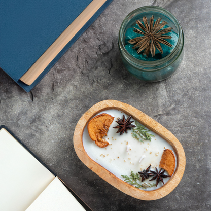 Candle with natural elements in a wooden holder on a gray surface with a book and small jar.