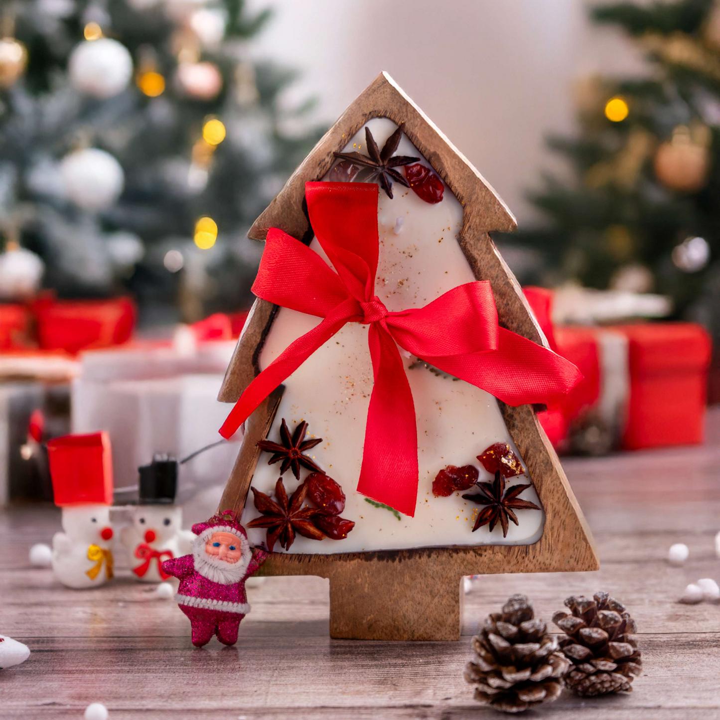 Decorative Christmas tree-shaped house with a red ribbon on a wooden surface, with Christmas trees and presents in the background.
