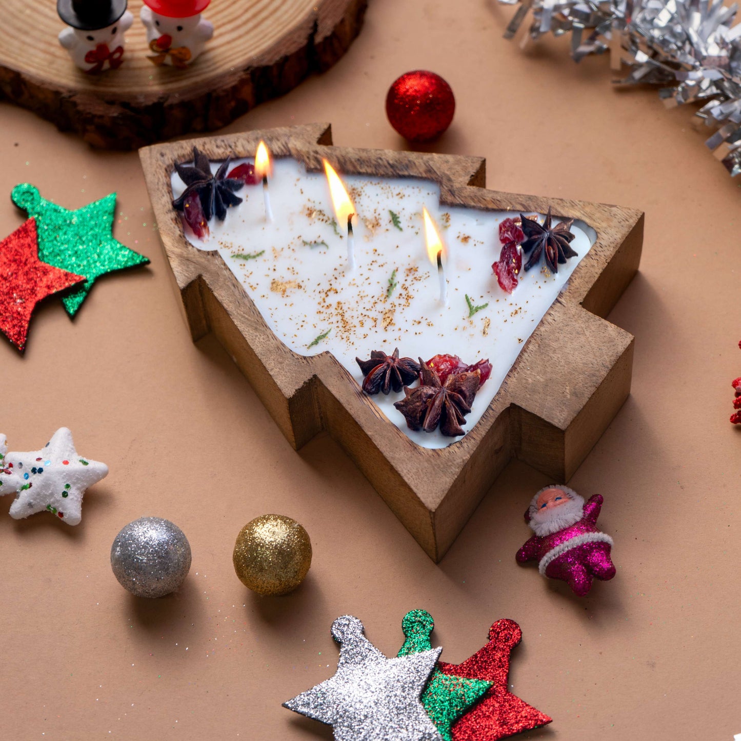 Decorative candle shaped like a Christmas tree with burning candles, surrounded by festive ornaments on a brown surface.
