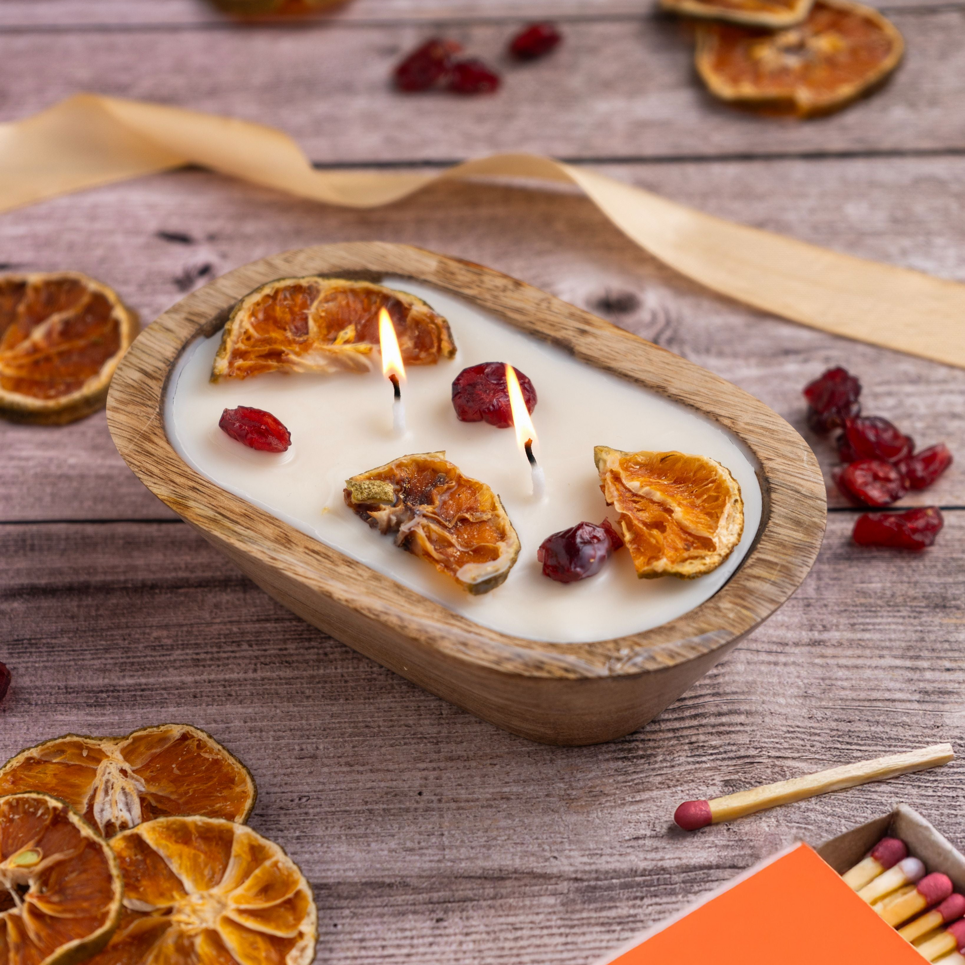 Candle in a wooden bowl with dried oranges and cranberries on a wooden surface, accompanied by matches.