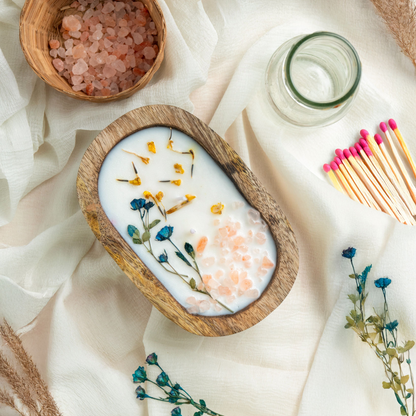 Wooden bowl with a candle featuring dried flowers, surrounded by a small bowl of pink salt, a glass jar, and matches on a white fabric background.