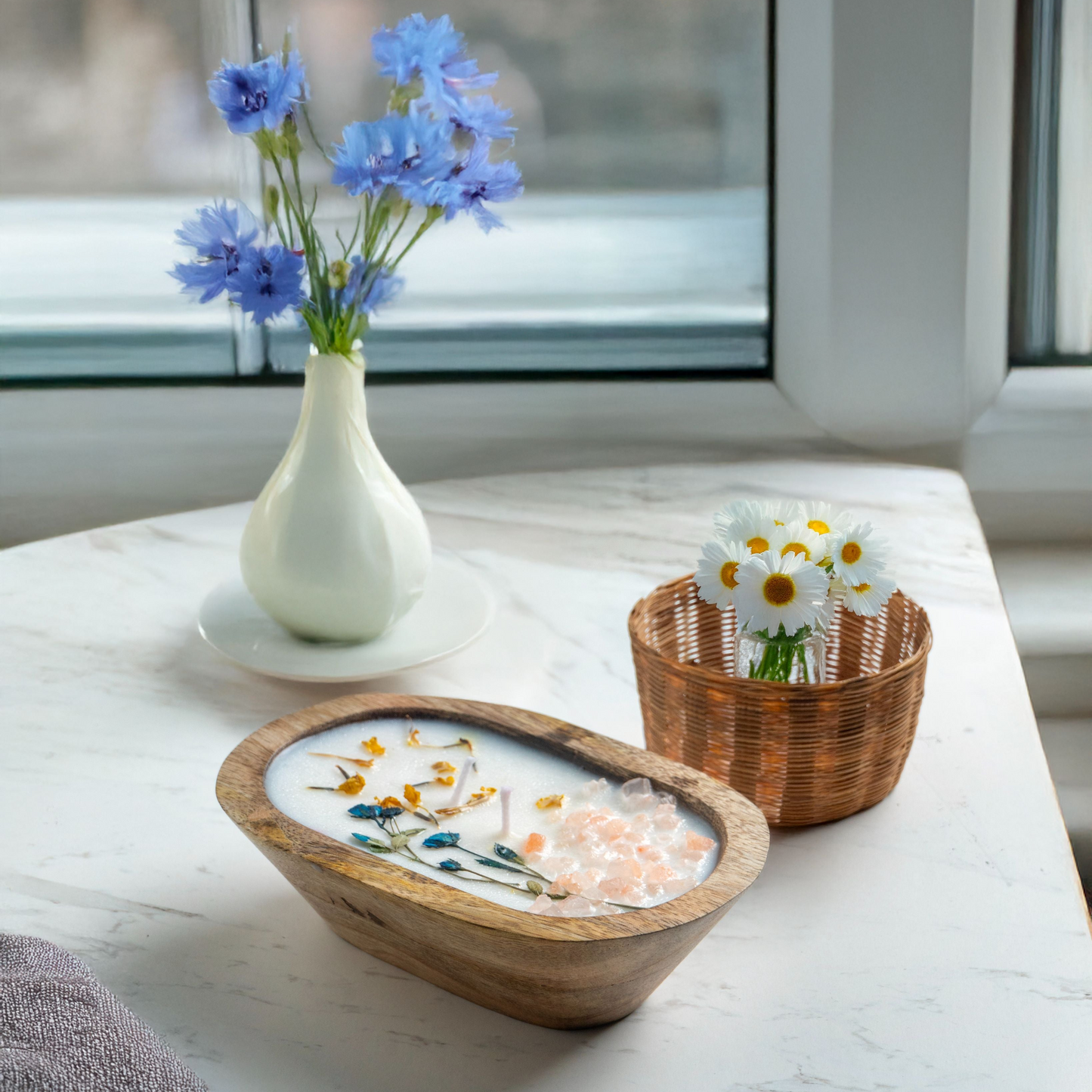 Decorative items including a vase with blue flowers, a bowl with decorative stones, and a basket with white flowers on a light surface.