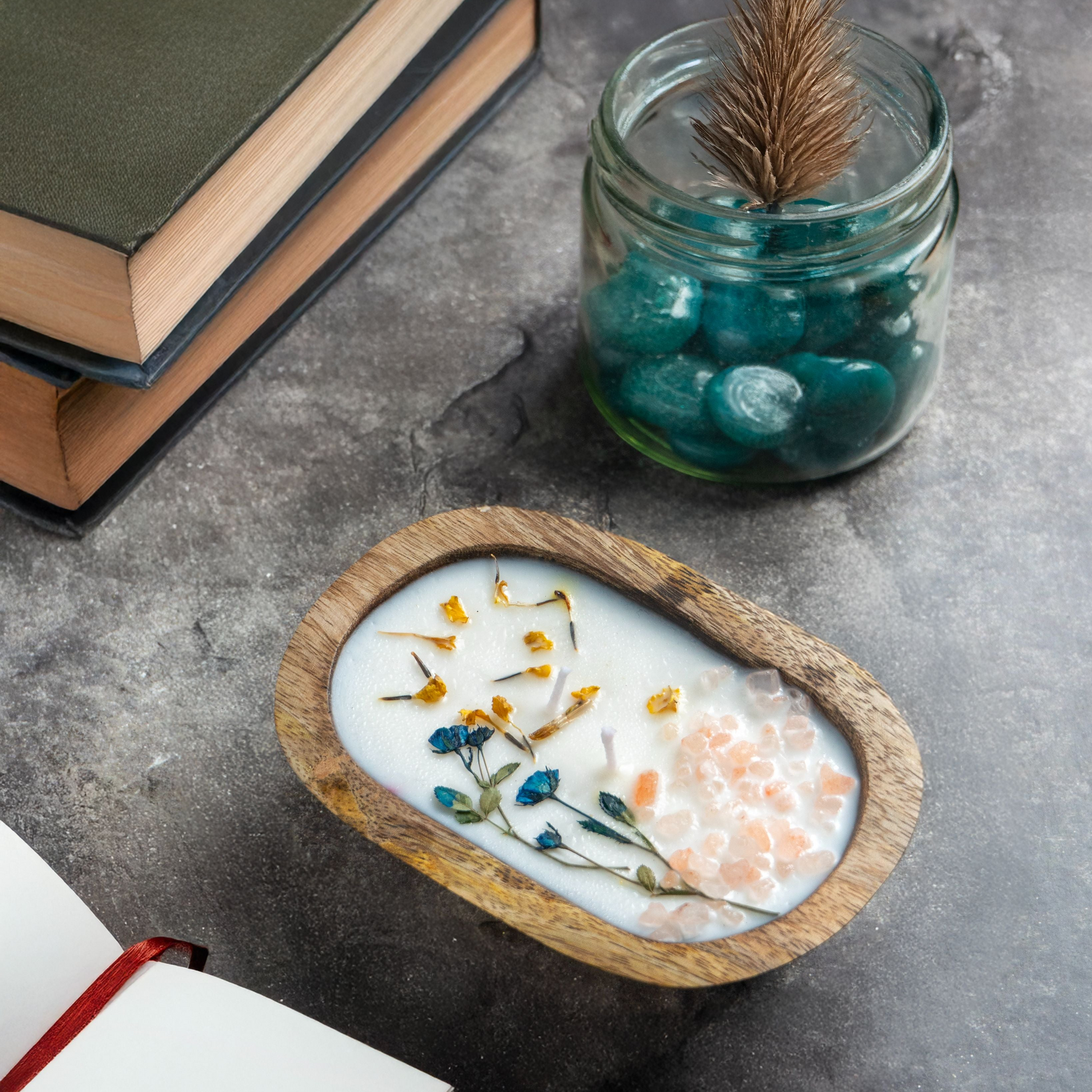 Candle in a wooden dish with flowers, books, and a jar on a concrete surface